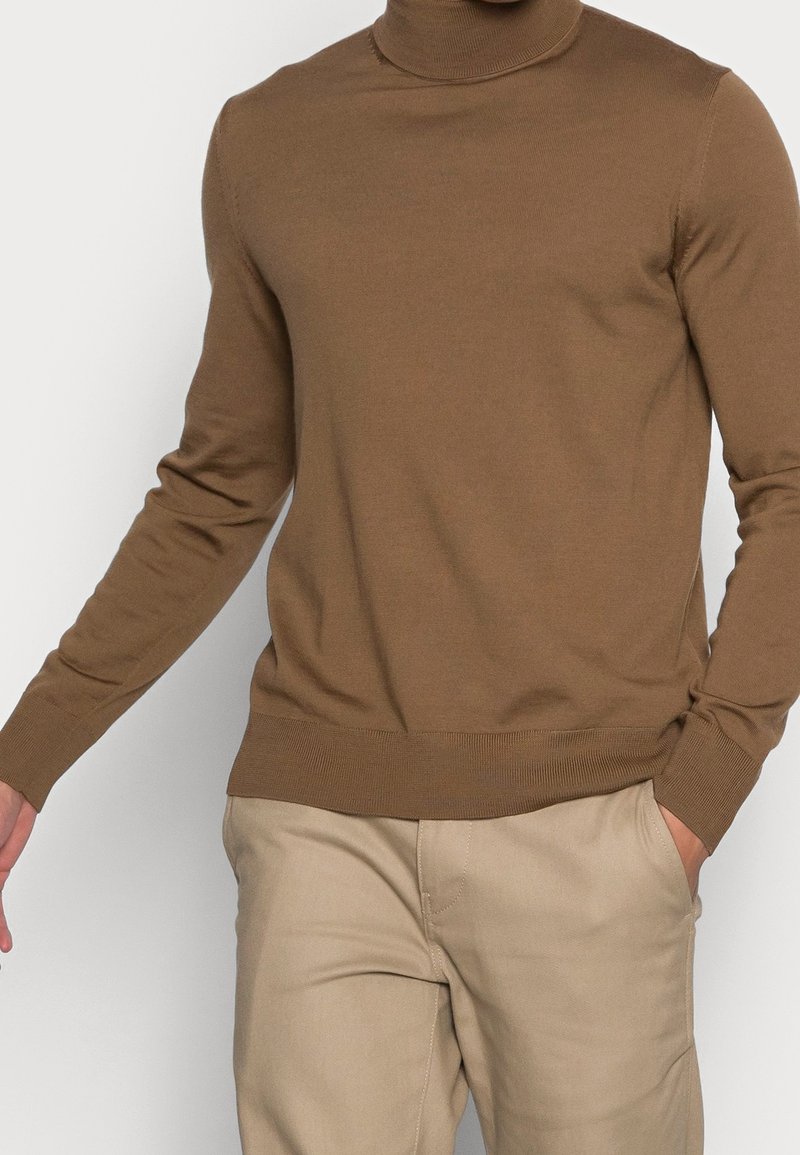 Brun polotröja i slät stickad tyg, med lång ärmar och ribbade muddar, parat med ljusbeiga byxor.