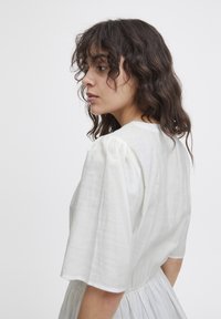 Robe blanche à manches bouffantes avec un bodice ajusté et une taille froncée, faite d'un tissu léger. Texture lisse et design simple.