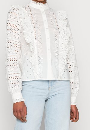 Kvinde iført en hvid bluse med hulmønster og flæser samt lyseblå jeans, stående mod en ensfarvet lys baggrund.