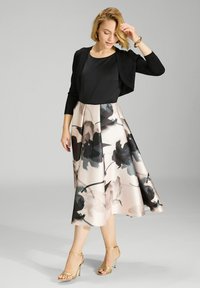 Chaqueta negra corta sobre una blusa negra, combinada con una falda de rosa claro hasta la rodilla que presenta un patrón floral negro y sandalias de tacón doradas.