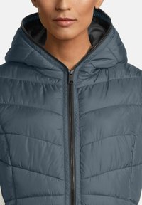 Vrouw in een donkerblauwe gewatteerde hoodie-jack, tot aan de nek dichtgeritst, zichtbaar vanaf de bovenste romp en een deel van het gezicht.