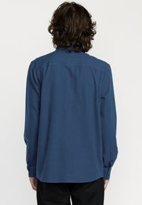 Chemise à manches longues en tissu bleu profond avec une texture lisse, dotée d'un poignet à boutons et d'un ourlet droit. La vue de dos montre le design du col.