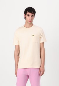 Krämfärgad bomullströja med bröstficka och en liten gul fjärilsikon. Modellen har på sig rosa shorts.
