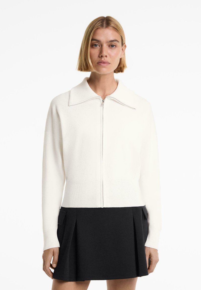 White zip-up cardigan with a large collar and ribbed hem, paired with a black pleated skirt. Soft fabric texture, minimalistic design.