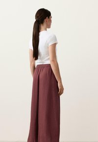 Femme aux longs cheveux bruns attachés en queue de cheval, portant une chemise blanche à manches courtes et un pantalon ample bordeaux taille haute, debout de profil.