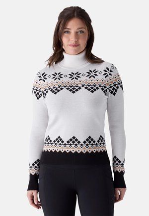Femme portant un pull à col roulé blanc avec des motifs géométriques et flocons noirs et beiges, associé à un pantalon noir.