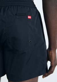 Black swim shorts made of lightweight material with an elastic waistband, featuring a back pocket and red Diesel logo label.