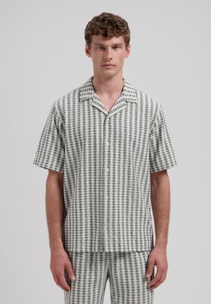 Mand iført kortærmet skjorte med knapper og matchende shorts med sort-hvid ternet teksturmønster, stående mod en ensfarvet baggrund.