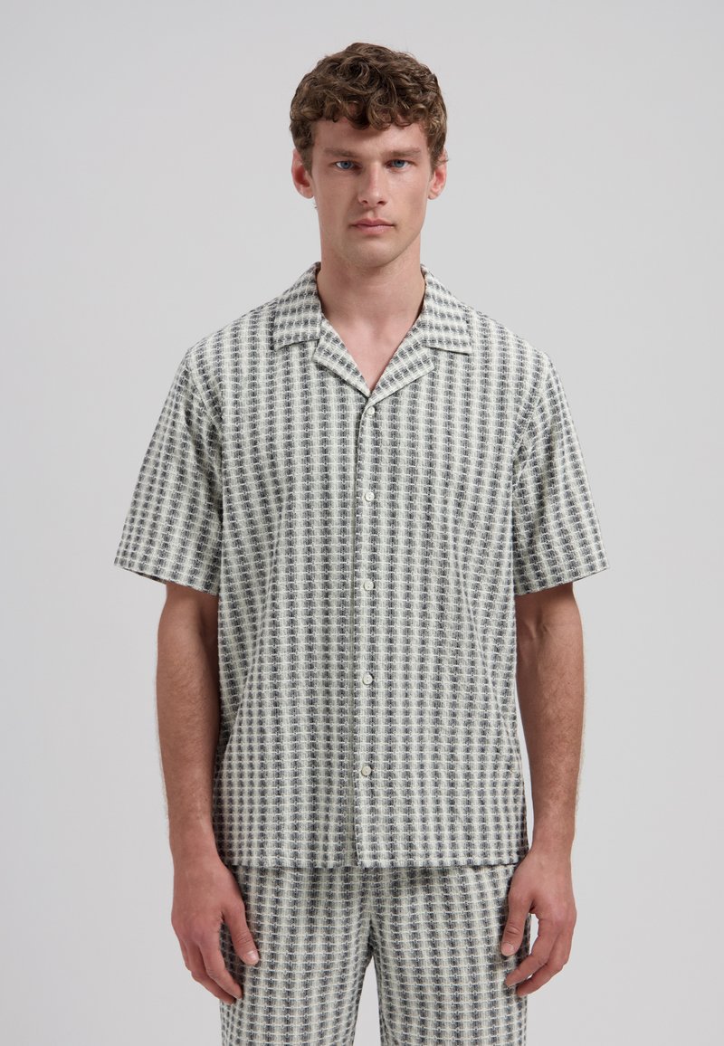 Homme portant une chemise à manches courtes boutonnée et un short assorti avec un motif texturé à carreaux noir et blanc, debout devant un fond uni.