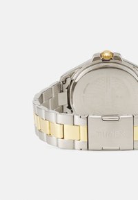 Montre en acier inoxydable argent et doré, avec un bracelet en métal bi-ton, une couronne texturée et une finition brossée à l'arrière du boîtier.