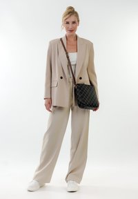 Beiger Blazer mit zwei schwarzen Knöpfen, cremefarbenem Rippoberteil, hellbeigen Hosen und einer schwarzen gesteppte Umhängetasche mit braunem Tragegurt. Weiße Sneakers.