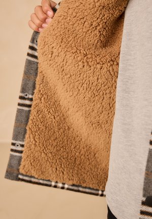 Main tenant une veste à carreaux grise ouverte, montrant sa doublure en polaire épaisse et douce de couleur tan sur un fond neutre.