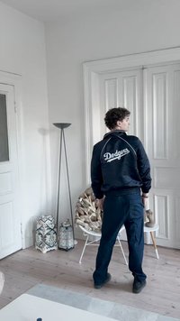 Fekete, selyem Dodgers dzseki fehér felirattal, sötétkék nadrággal kombinálva. A szoba fehér falakkal, fa padlóval és dekoratív tárgyakkal rendelkezik.