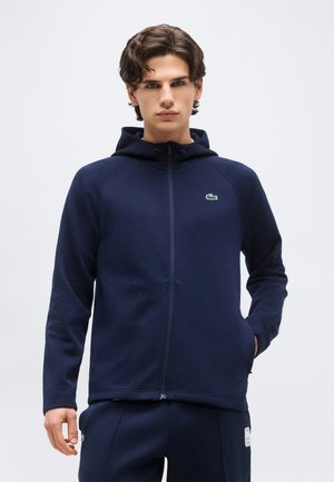 Νέος άνδρας με κοντά σκούρα μαλλιά, φορώντας ένα μπλε-σκούρο φούτερ με φερμουάρ και ένα μικρό πράσινο λογότυπο στο στήθος, στέκεται μπροστά σε ένα απλό φόντο.