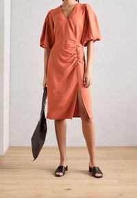 Robe couleur rouille avec des manches bouffantes, décolleté en v, détails froncés sur les côtés et fente haute sur la cuisse. Associée à des sandales à talons noires et un sac à main.