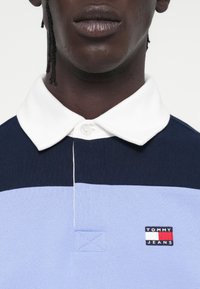 Koszulka polo z kołnierzykiem w kolorze granatowym, jasnoniebieskim i białym w blokach kolorystycznych. Posiada haftowane logo "TOMMY JEANS" oraz teksturowany materiał.