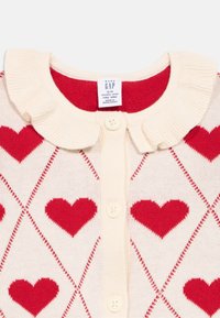 Krémový kardigan s červeným vzorem srdce a volánkovým límcem. Zapínání na tři knoflíky. Jemná pletená textura.