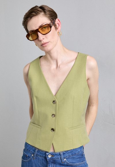 Chaleco de lino verde con un profundo escote en V, que cuenta con dos bolsillos frontales y botones marrones. Combinado con jeans de mezclilla azul y gafas de sol de tonalidad marrón.