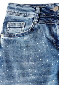 Jeans de denim azul con un acabado texturizado, con tachuelas metálicas distribuidas de manera uniforme por toda la tela. Incluye un bolsillo frontal y cierre de botón.