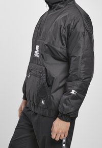 Black pullover jacket made of shiny nylon, features a zippered front, large front pocket, elastic cuffs, and a logo on the chest and sleeve.