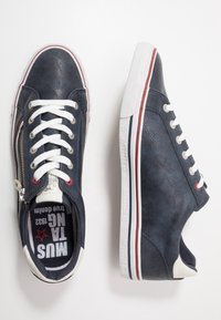 Baskets en cuir bleu marine avec lacets blancs, fermeture éclair latérale et bandes contrastantes rouges et blanches sur la semelle. Finition texturée et détail du logo.