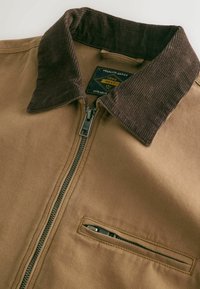 Veste marron avec col en velours côtelé, fermeture éclair centrale et poche poitrine zippée, avec une étiquette indiquant "Premium Grade 1982 Next Durable".