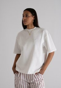 White cotton t-shirt with short sleeves and a relaxed fit. Features a subtle embroidered logo on the chest, paired with striped linen pants.