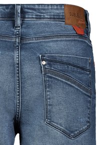 Rückseite einer blauen Jeans aus Denim mit einer detaillierten Tasche, braunem Lederetikett der Marke SUBLEVEL und einer orangefarbenen Stoffschlaufe am Bund.