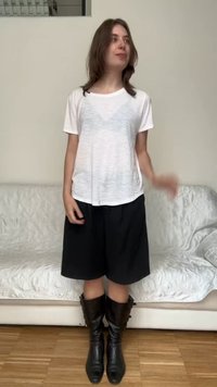 Jeune femme debout à l'intérieur portant un t-shirt blanc, une jupe noire longueur genou et des bottes en cuir noires devant un canapé blanc.