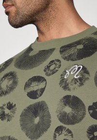 Πράσινο ελιάς T-shirt με μαύρο κυκλικό μοτίβο και λευκό κεντημένο λογότυπο στο στήθος. Κατασκευασμένο από μαλακό βαμβακερό ύφασμα.