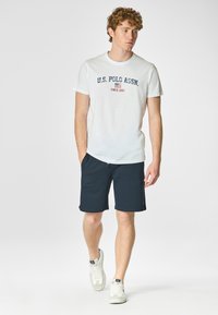 T-shirt bianco a maniche corte con scritta blu "U.S. POLO ASSN." e accento rosso, abbinato a pantaloni corti blu scuro e sneakers bianche.
