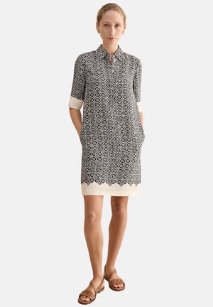 Mujer de pie con las manos en los bolsillos, lleva un vestido hasta la rodilla con patrón en blanco y negro y sandalias planas marrones sobre un fondo liso.