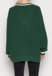 Pull vert en tricot à coupe ample, ourlet côtelé, et détail de rayure blanche autour du col et des poignets. Tissu texturé avec manches longues.