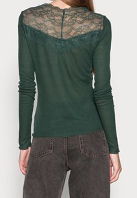 Zelený dlouhý rukáv top s krajkovým detailem na zadní straně, s kulatým výstřihem a přiléhavými rukávy. Materiál se zdá být měkký a mírně elastický.