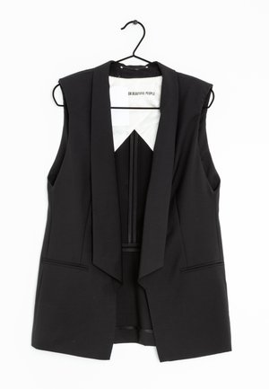 Chaleco blazer negro sin mangas, entallado, con solapas, colgado en una percha negra sobre fondo blanco.