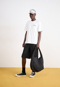 White cotton t-shirt with a logo, black shorts, black quilted tote bag, and black sneakers with white soles, set against a yellow background.