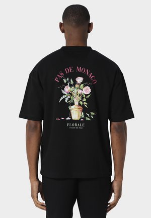 Uomo che indossa una maglietta nera con un'illustrazione di un vaso floreale e la scritta "Pas de Monaco" e "Florale à fleur de peau" sulla schiena.