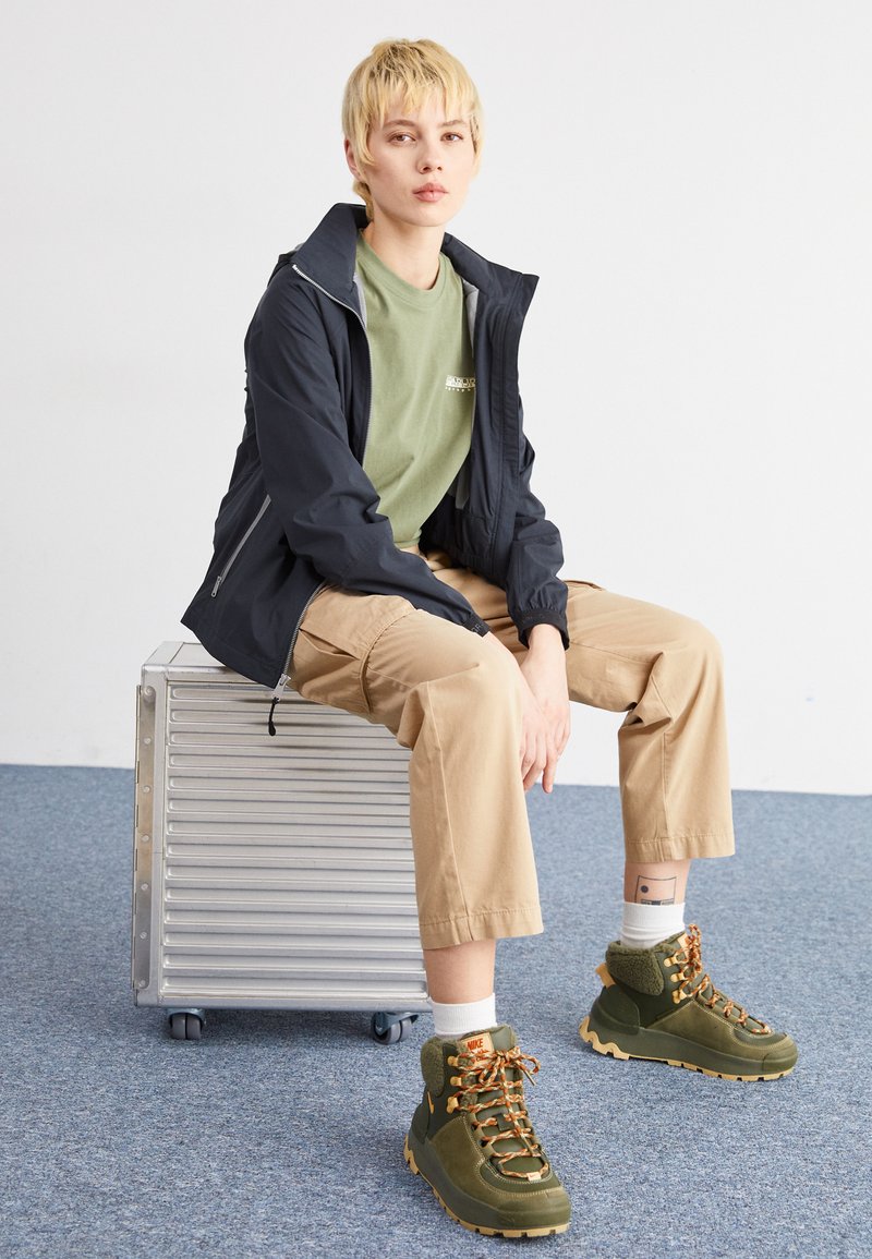 Giacca nera leggera, t-shirt verde oliva, pantaloni cargo beige e scarpe da trekking verdi con lacci arancioni. Valigia metallica su un tappeto blu.