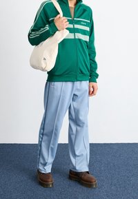 Grön och ljusblå träningsoverall med tre vita ränder, zip-jacka och lösa byxor. Canvasväska i handen, bruna stövlar på fötterna.