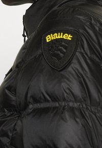 Czarna kurtka puchowa z pikowaną teksturą, z wyraźnym, okrągłym czarno-żółtym logo na lewym ramieniu.