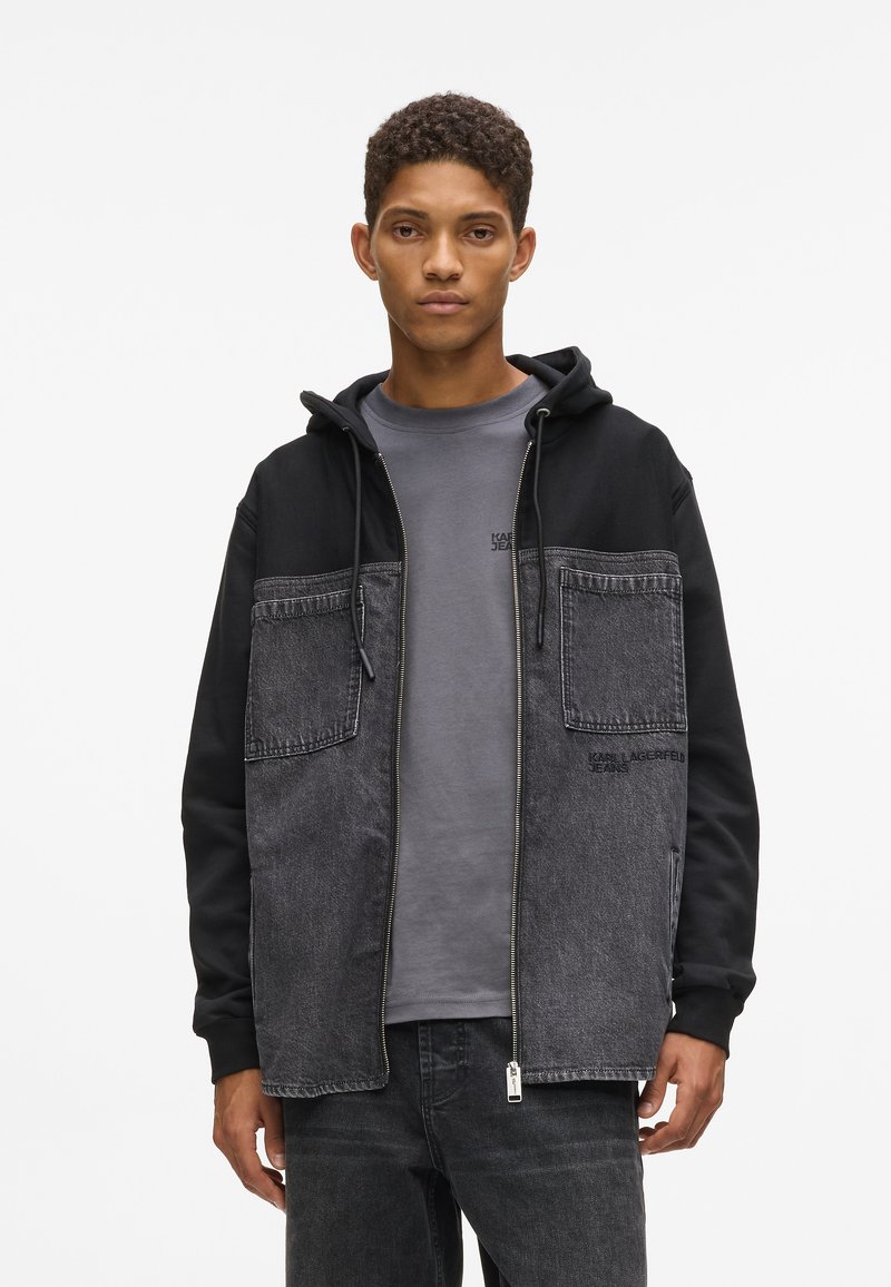 Μαύρο και γκρι μπουφάν με φερμουάρ μπροστά, που διαθέτει δύο μεγάλες πλαϊνές τσέπες, κουκούλα και ένα μείγμα αντίθετων υφασμάτων. Συνδυάζεται με ένα γκρι πουκάμισο.