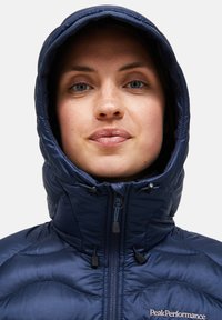 Tmavě modrá izolovaná bunda s vysokým límcem a připojenou kapucí. Má zipové zapínání a hladkou prošívanou texturu. Na hrudi je logo.