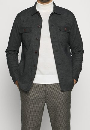 Mand iført mørkegrå skjortejakke med knapper over hvid rullekrave og ternede brune og beige bukser med hænderne i lommerne.