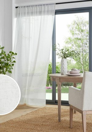 Una tenda bianca trasparente copre parte della porta a vetri con un tavolo in legno chiaro, una sedia, un vaso con piante verdi e un giardino verde all'esterno.