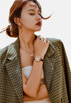 Reloj de tono dorado con esfera redonda, correa de cuero negro y acentos de cristal. El modelo lleva un blazer de cuadros verdes y un delicado collar de oro.