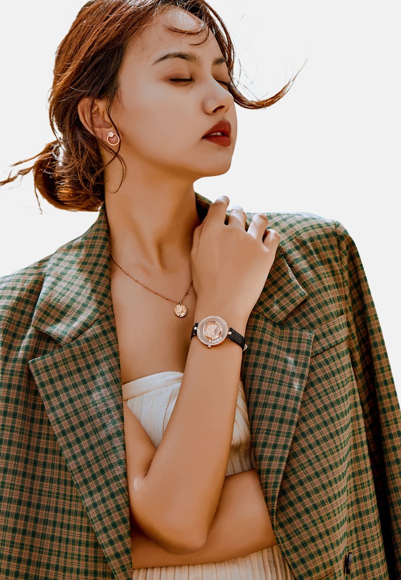 Reloj de tono dorado con esfera redonda, correa de cuero negro y acentos de cristal. El modelo lleva un blazer de cuadros verdes y un delicado collar de oro.