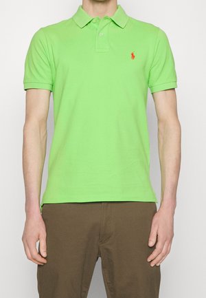 T-shirt polo en coton vert avec un col, des manches courtes et un petit logo rouge sur la poitrine. Porté avec un pantalon marron.