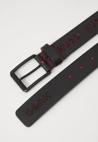 Ceinture en cuir noir avec logo "HUGO" embossé, surpiqûres rouges et boucle en métal noir. Elle dispose de plusieurs trous d'ajustement le long de la sangle.