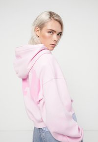 Sudadera de forro polar rosa con bolsillo tipo canguro, que presenta un gran logo rosa en la parte posterior. El modelo la lleva con jeans de un azul claro.