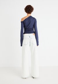 Top bleumarin cu un umăr, cu textură plisată, asortat cu blugi albi, cu talie înaltă și picioare late, prezentat din spate. Design curat, minimalist.